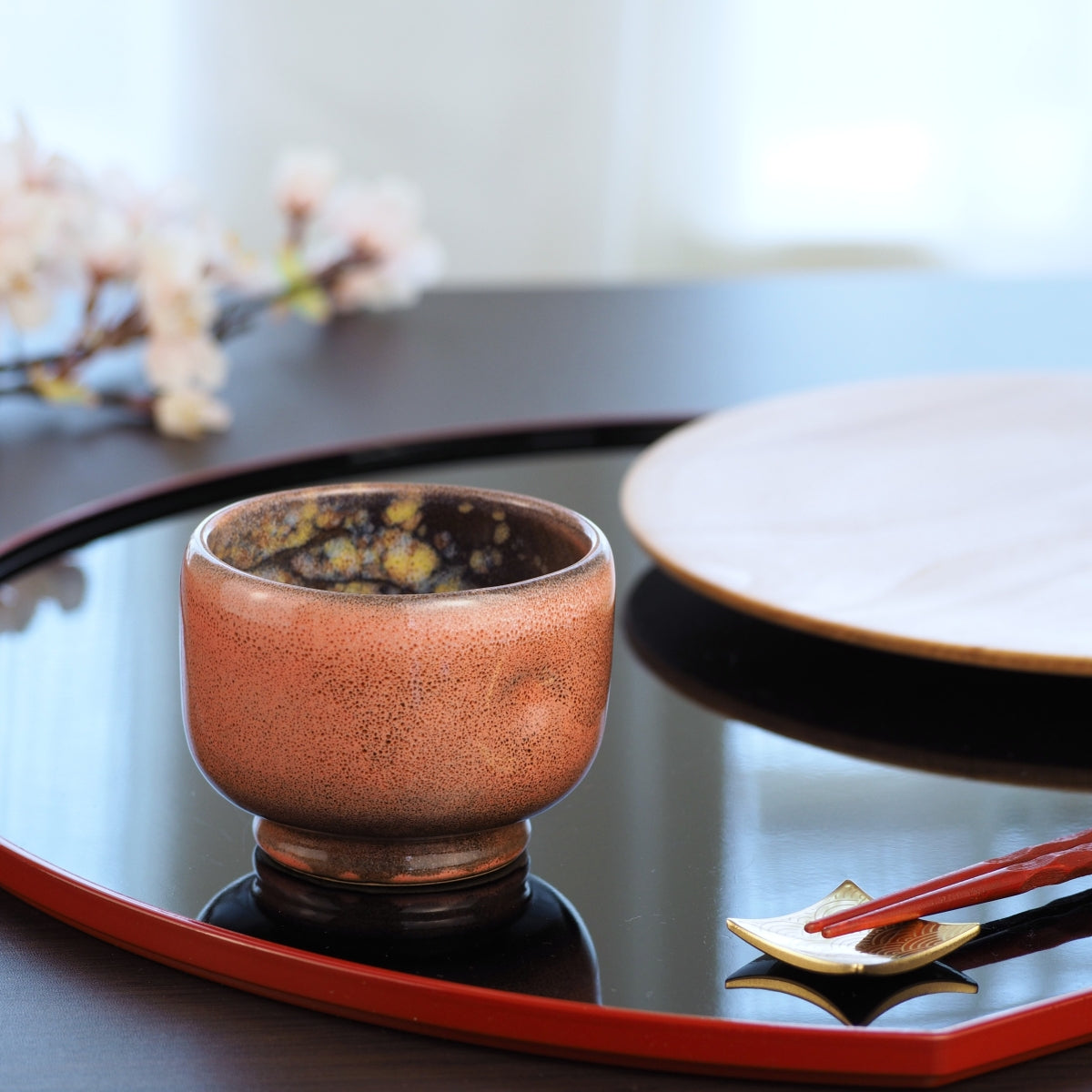 A Japanese ceramic teacup on a reflective surface with a blurred background in a lifestyle setting. Tsukushi