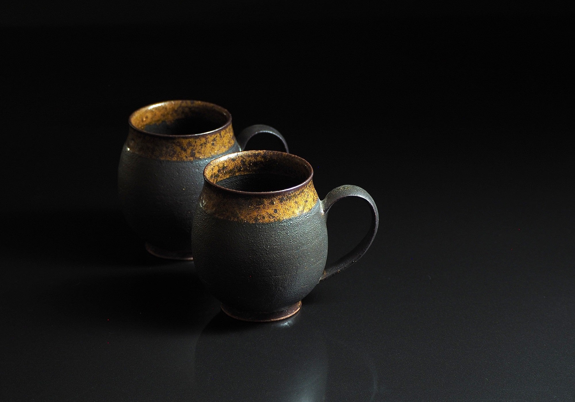 Handcrafted two black Japanese ceramic tea mugs with a rustic finish on a black background, Tsukushi