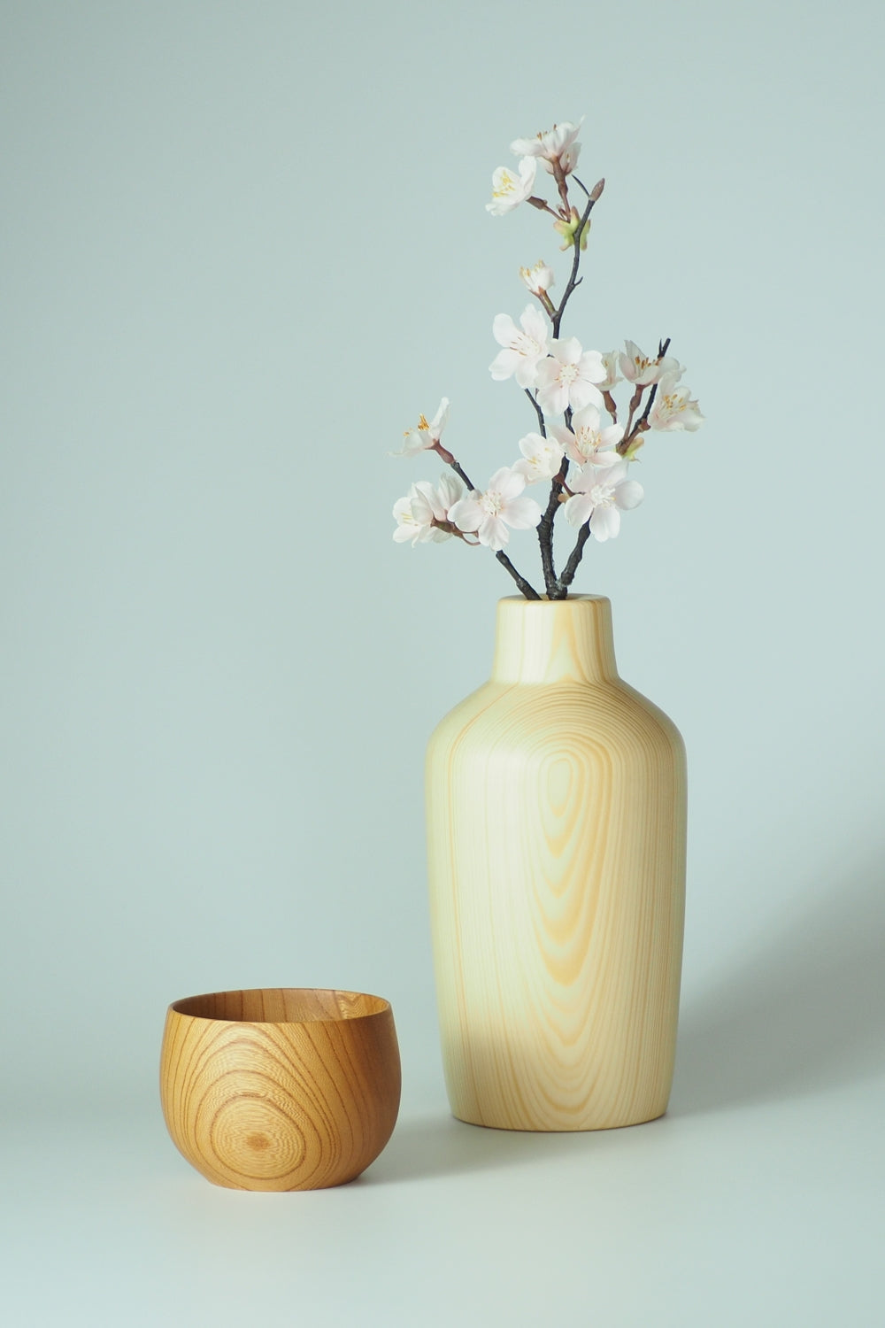 A wooden vase with cherry blossoms and a matching wooden cup on a light gray background. Tsukushi