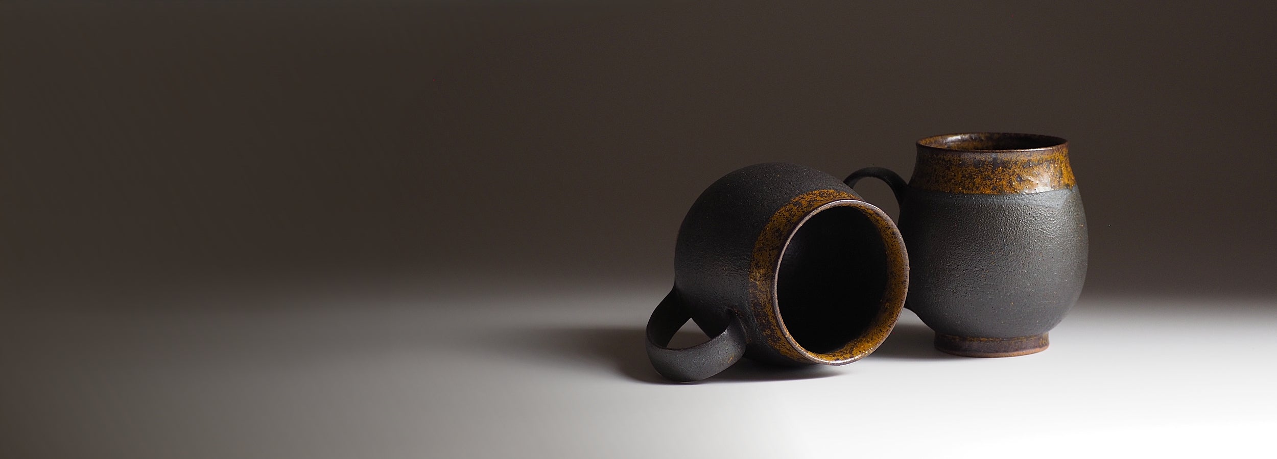Two handcrafted Japanese ceramic mugs on a gray surface with a dark background. Tsukushi
