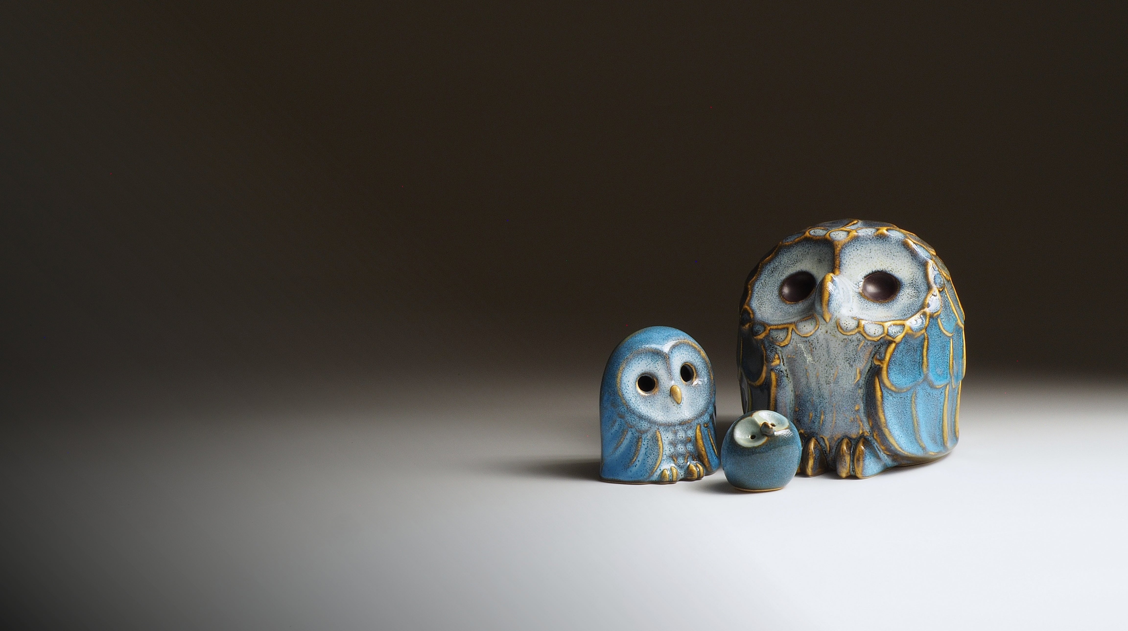 Three handmade ceramic owl figurines in shades of blue, gray, and gold, displayed on a white surface against a dark brown background.