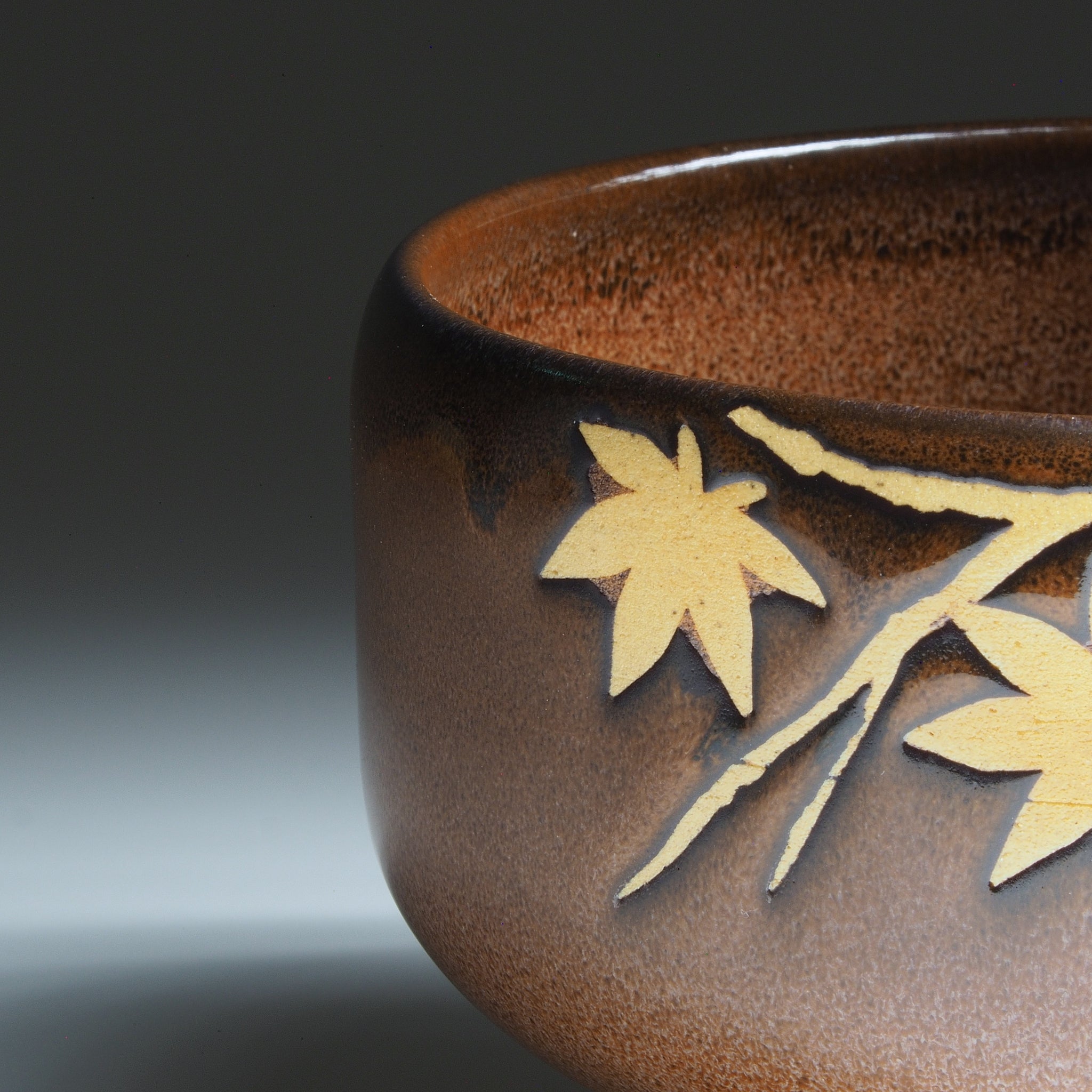 Close-up of Yūkaede matcha bowl, momiji maple leaves in natural clay against Nanasai Tenmoku brown glaze