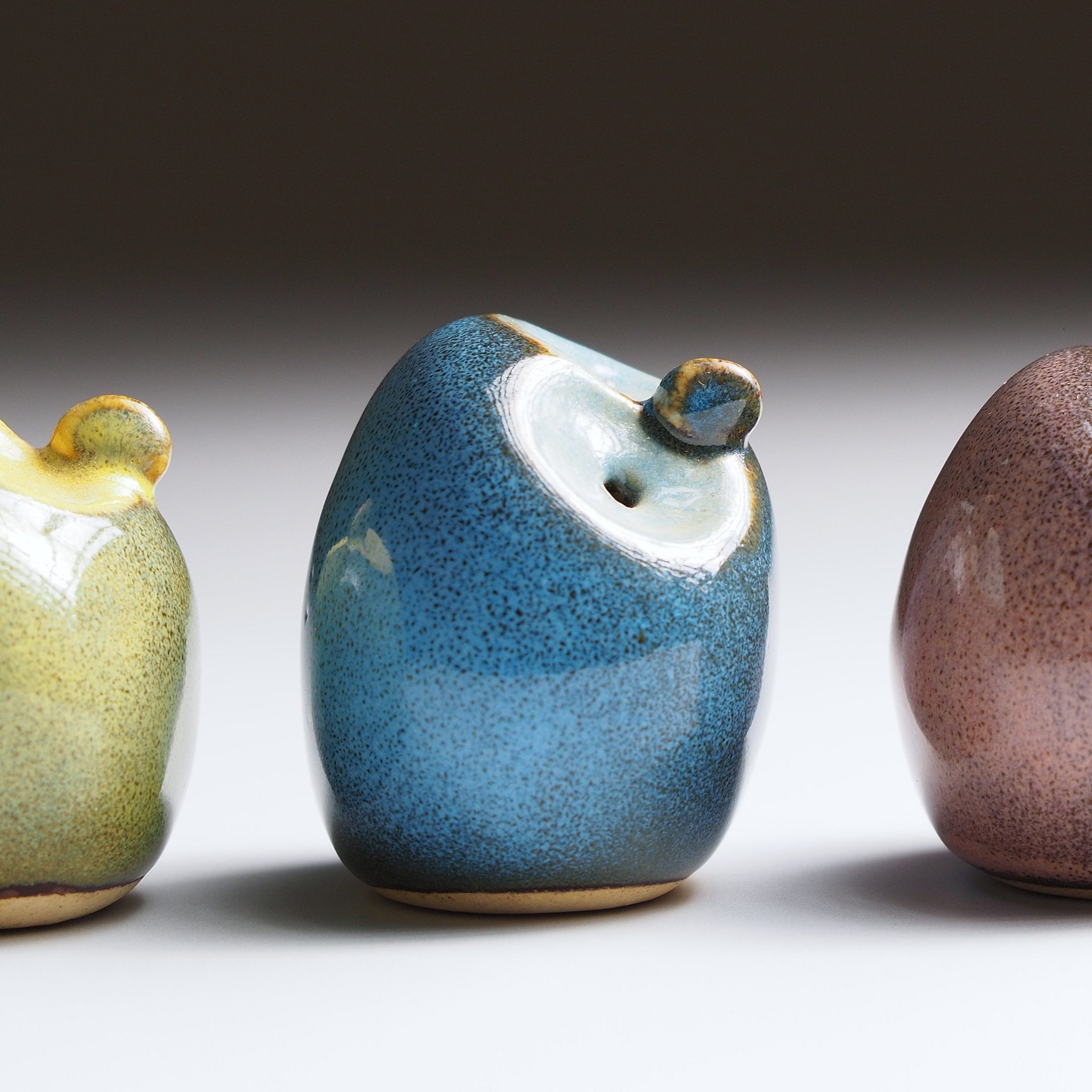 Gofuku ceramic owl figurines in lineup detail showing moss chartreuse, twilight blue, and dusk plum glazes with gold beak features.