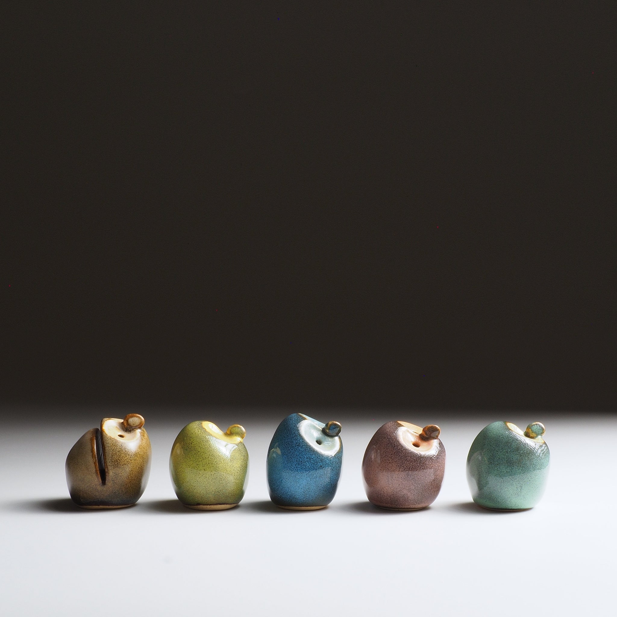 Five Gofuku ceramic owl figurines arranged in a line showing all glaze colors: sandstone, moss chartreuse, twilight blue, dusk plum, and misty celadon.