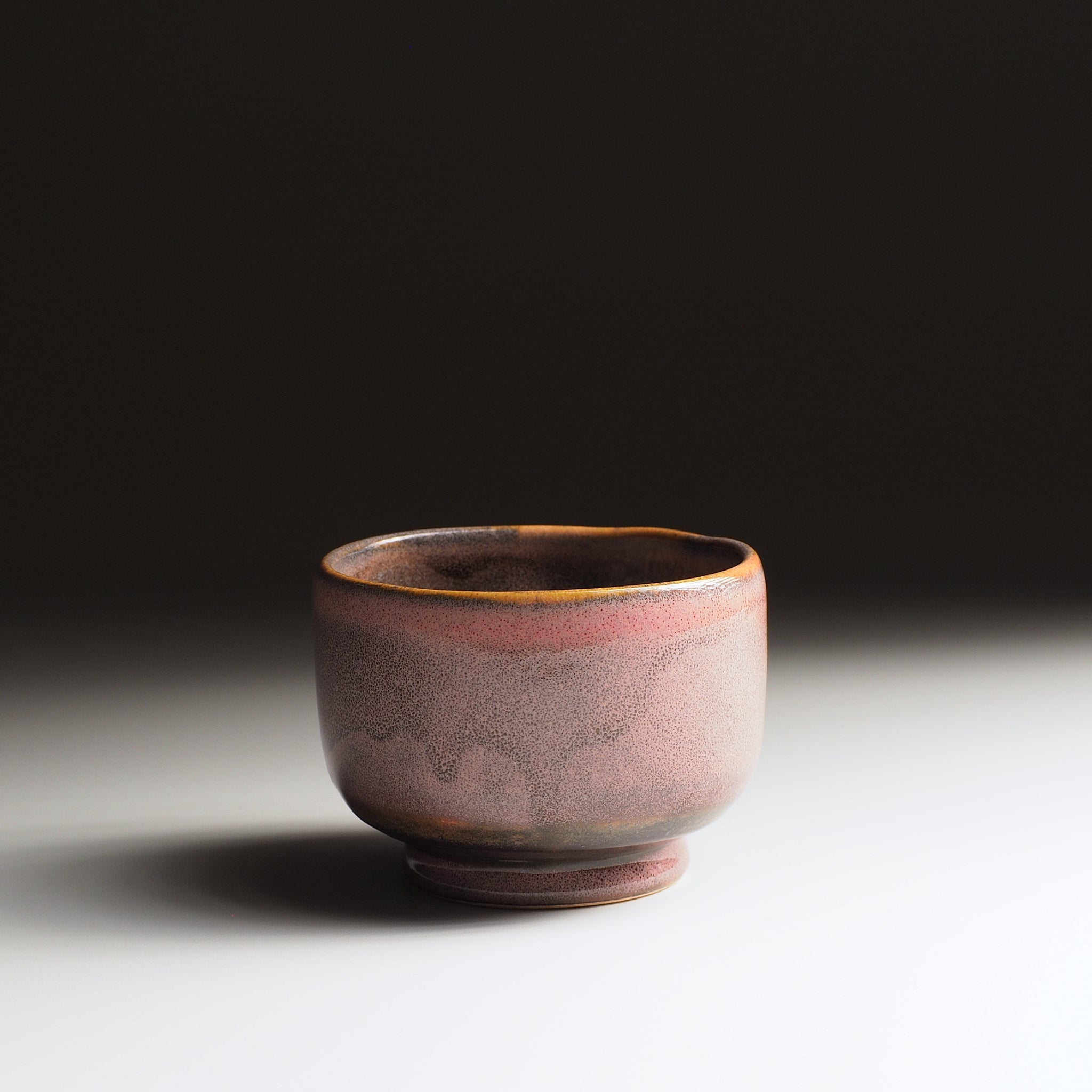 Handmade round teacup with a speckled pink and brown glaze and golden rim, on a dark-to-light gradient background.